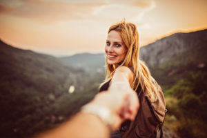 Girl is holding hand of her man high on mountain in sunset.